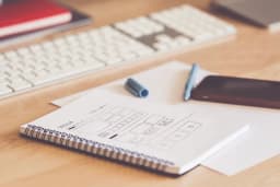 A spiral notebook on a desk showing a hand-drawn website layout sketch, beside a white keyboard, a pen and a phone