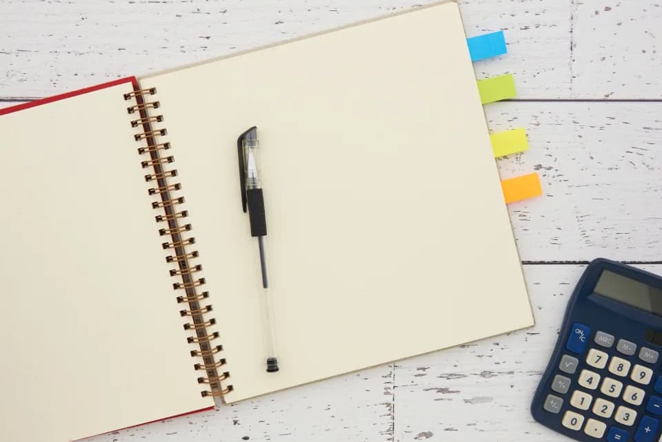 An open spiral notebook with blank cream pages and a black pen, next to a blue pocket calculator and coloured sticky tabs on a white painted wooden desk