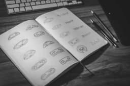 An open sketchbook filled with hand-drawn robot logo concepts and lettering, next to pencils, a keyboard and a graphics tablet