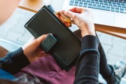 A person taking a bank card out of a black leather wallet while sitting in front of an open laptop