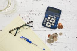 Overhead view of a blue desk calculator, US coins, a yellow accounting ledger pad, reading glasses and a blue pen on white painted wooden boards