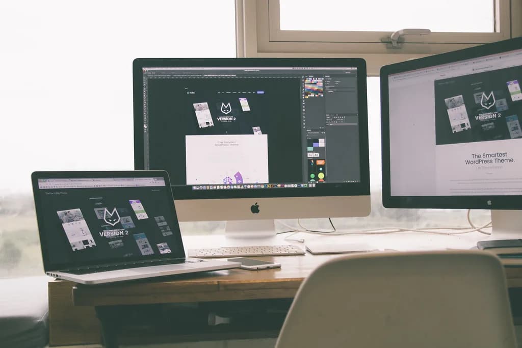 A designer's desk with a laptop and two monitors all showing the same website design at different screen sizes