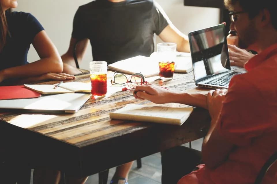 Four people working around a reclaimed wood table with an open laptop, notebooks and iced drinks in a bright room