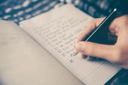 A hand writing a checklist with empty tick boxes in a grid-paper notebook using a black pen