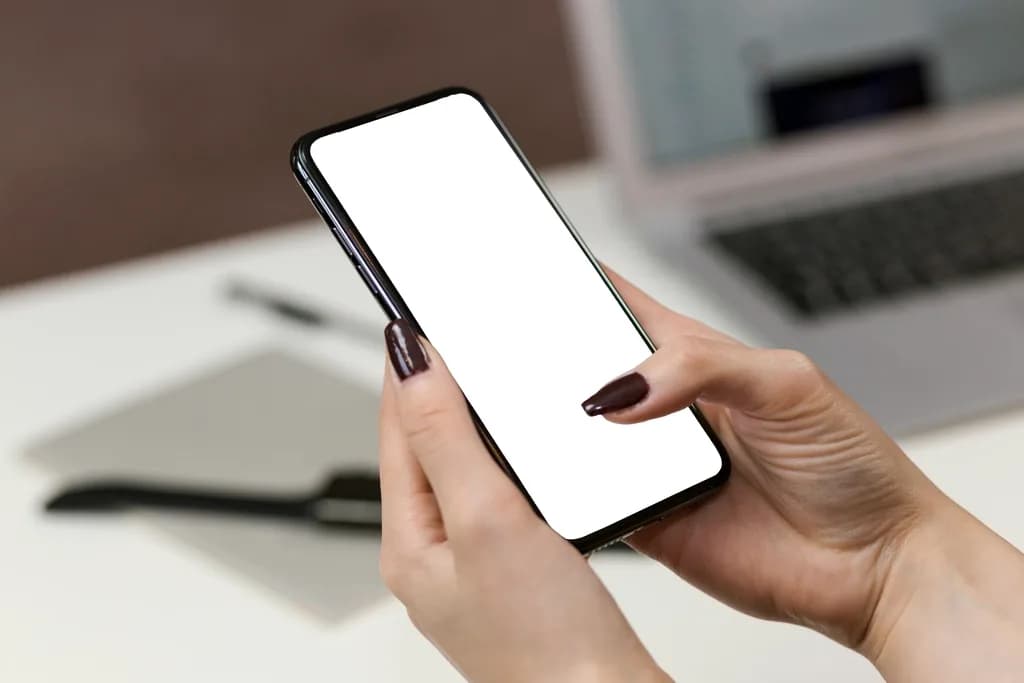 A person holding a smartphone with a blank white screen in both hands, with a laptop out of focus on the desk behind