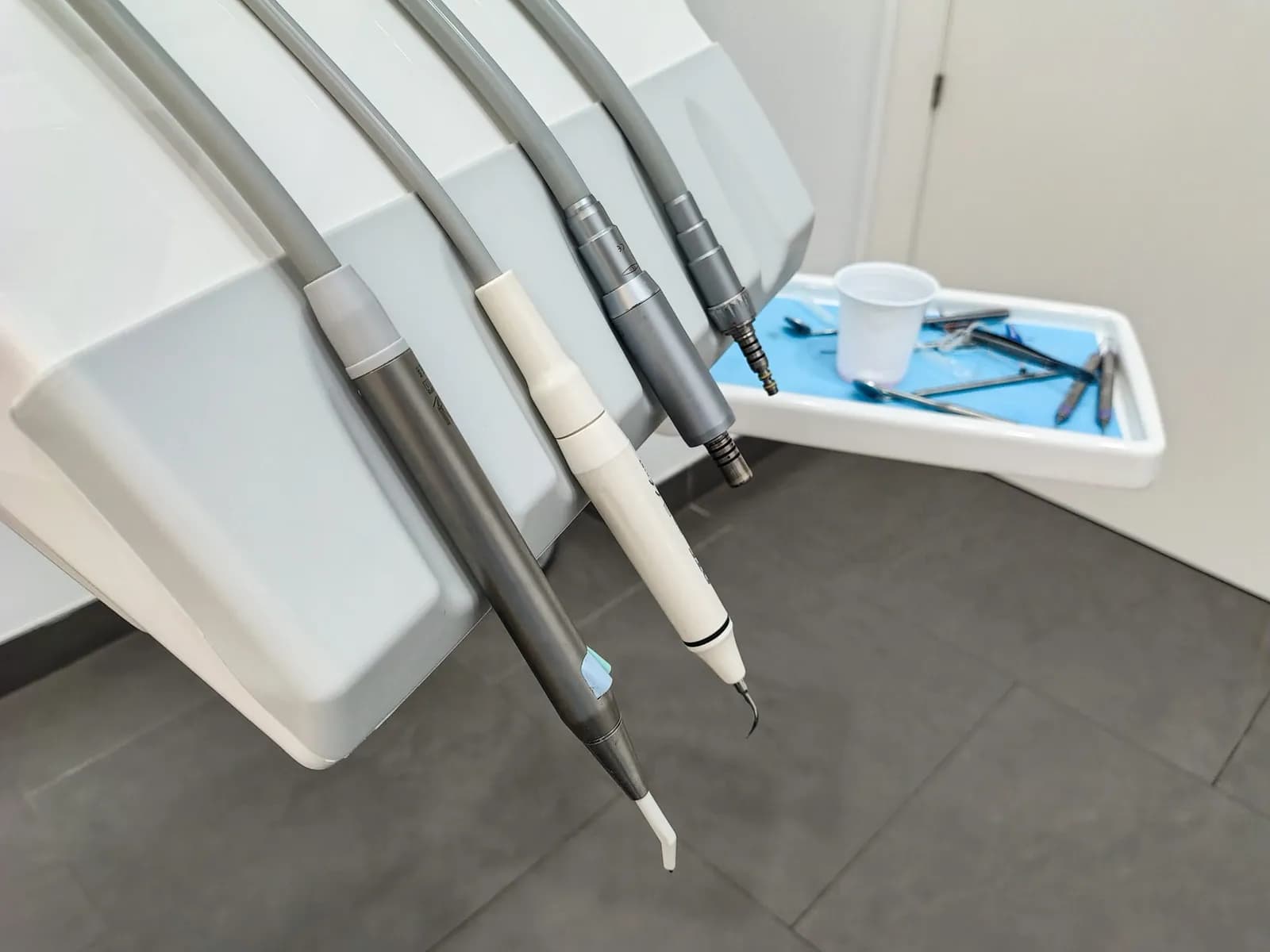 Dental handpieces hanging from a delivery unit beside a tray of instruments and a plastic rinsing cup