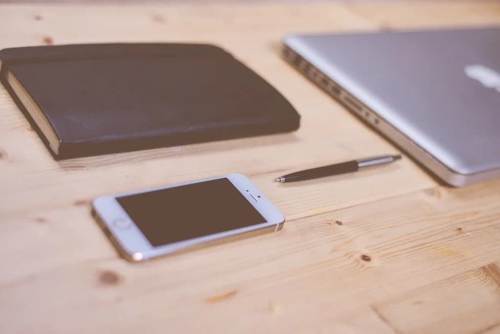 A white smartphone with a blank screen, a closed leather notebook, a pen and a closed silver laptop arranged on a pale wooden desk