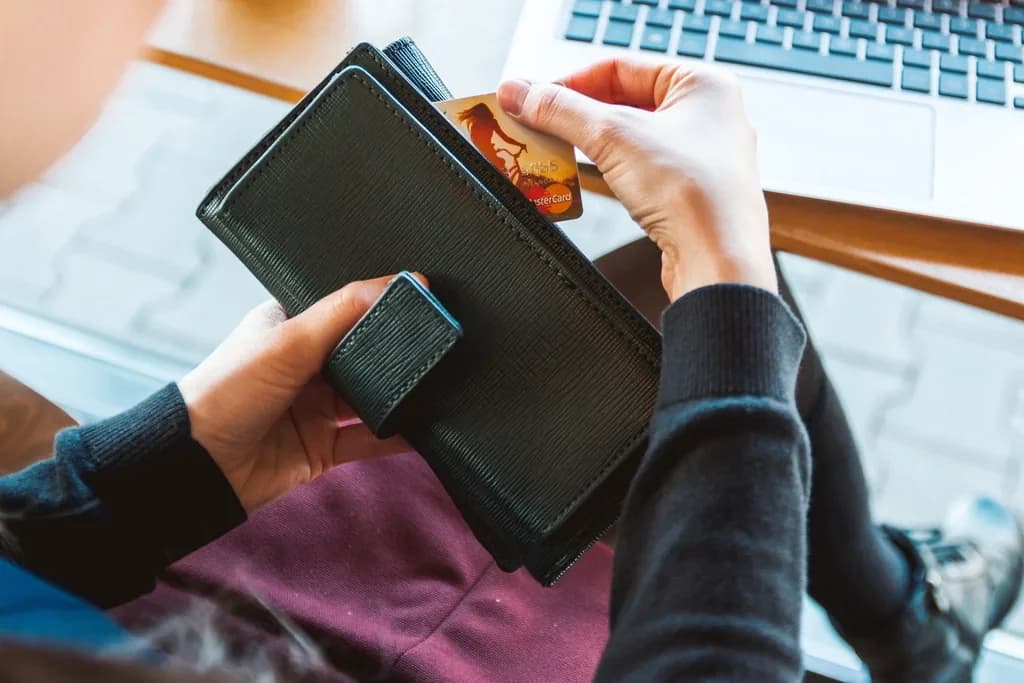 A person taking a bank card out of a black leather wallet while sitting in front of an open laptop