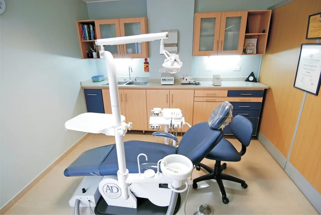 Empty dental treatment room with a blue dental chair, overhead examination lamp and framed certificates on the wall