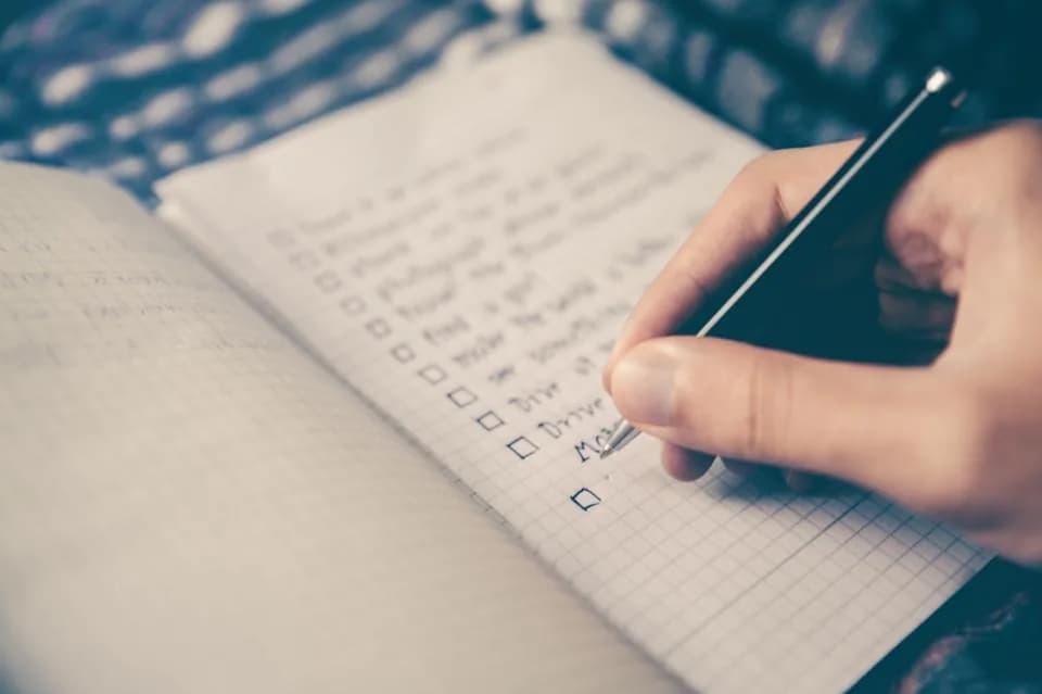 A hand writing a checklist with empty tick boxes in a grid-paper notebook using a black pen
