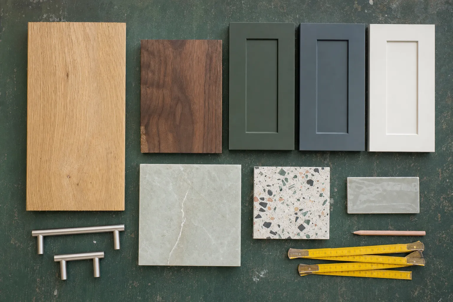 Samples on a green workbench: oak, walnut, painted doors in green, grey and white, quartzite, terrazzo, a green zellige tile, steel handles and a folding rule