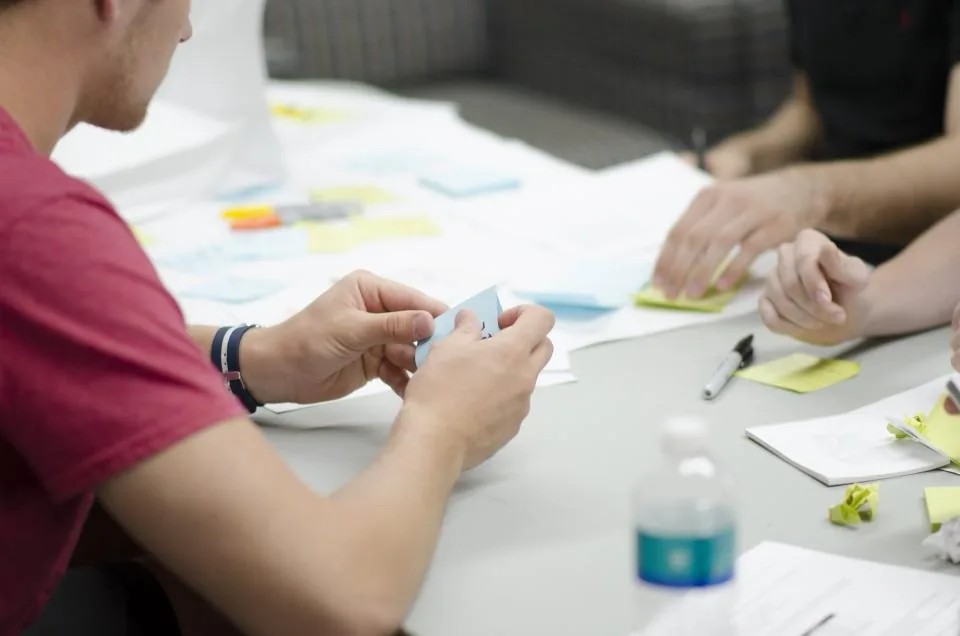 Three people seated around a table sorting blue and yellow sticky notes over printed sheets of paper