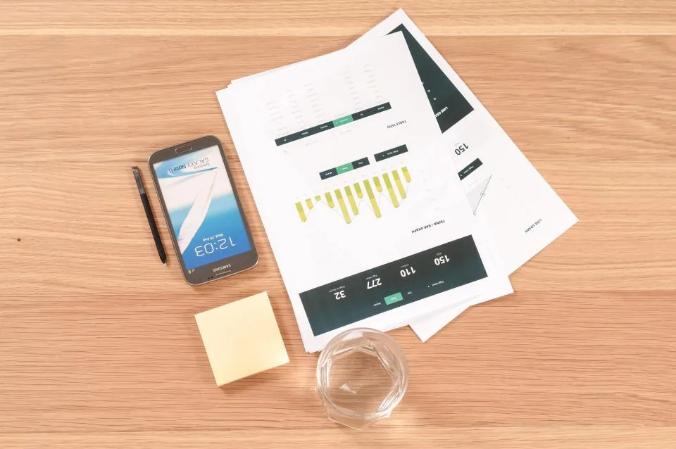 Printed report pages showing bar charts and data tables on a wooden desk beside a smartphone, a stylus and a yellow sticky note