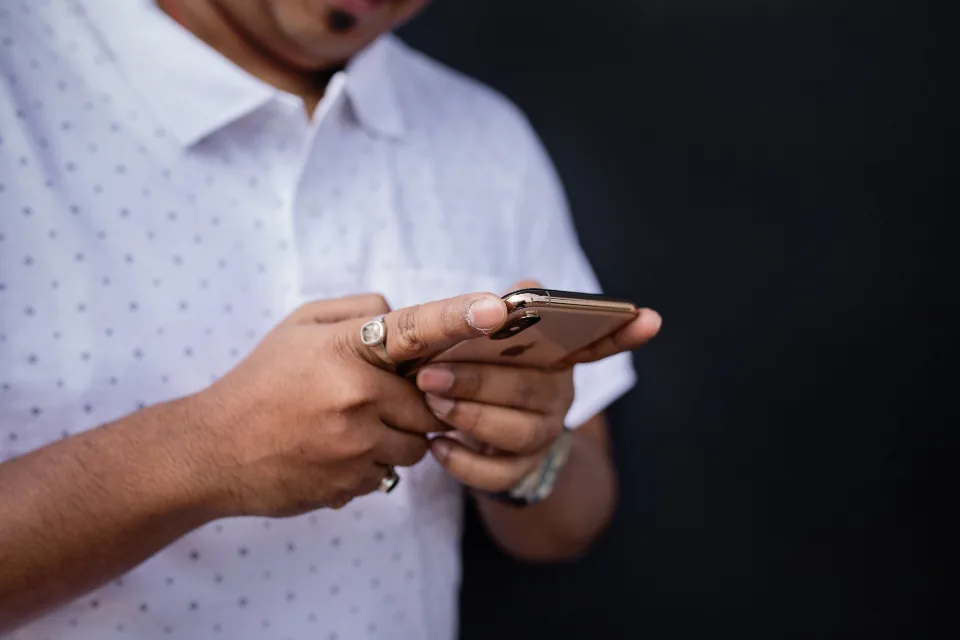 A man in a patterned white shirt typing on a smartphone held in both hands against a dark background