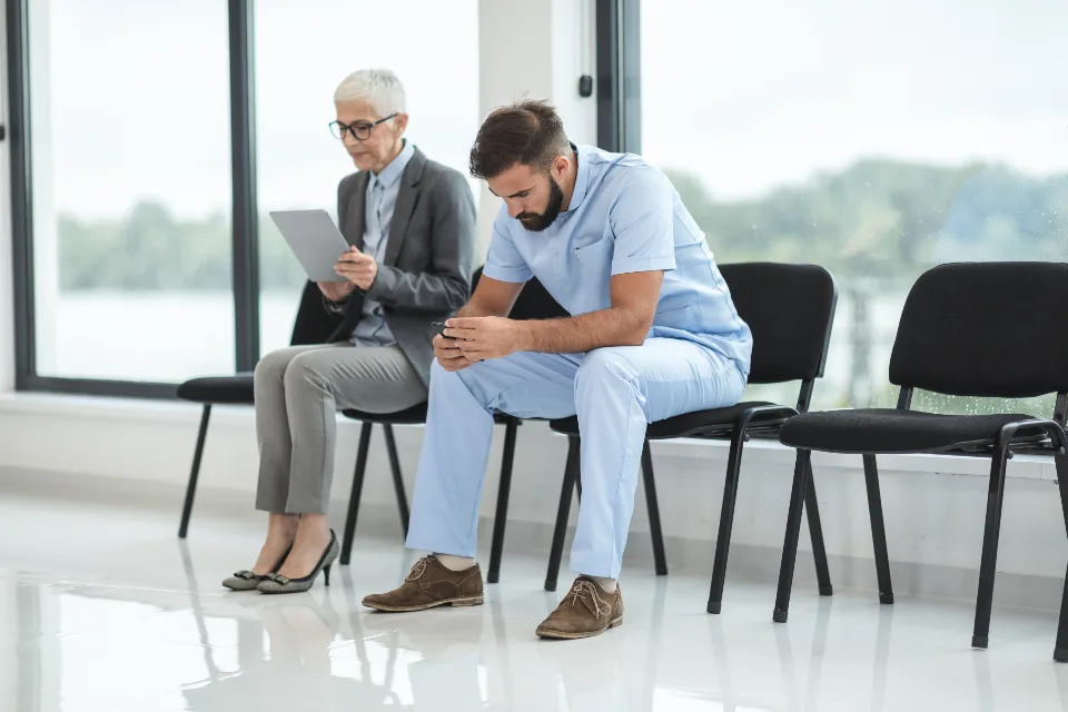 Two people sitting on black chairs in a bright waiting area beside tall windows, one in blue medical scrubs looking at a phone and one holding a tablet