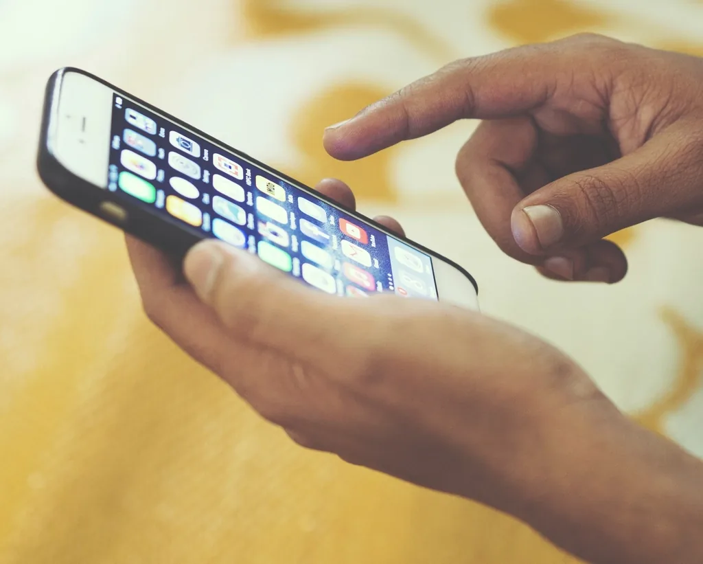 Two hands holding a smartphone that shows a home screen full of app icons, with one index finger about to tap the screen