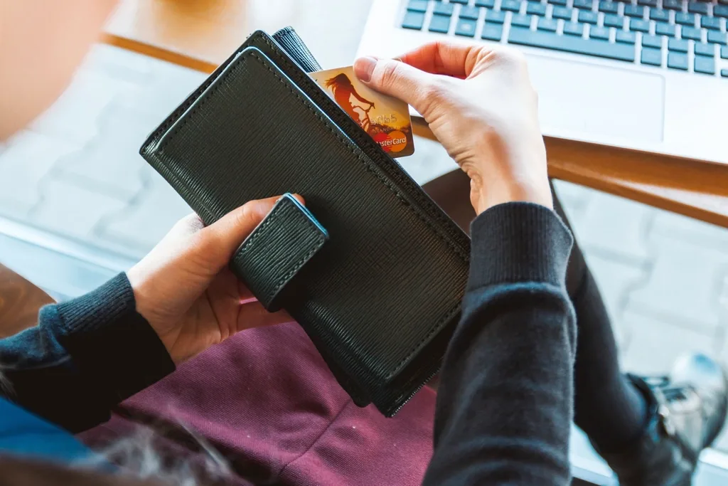 A person taking a bank card out of a black leather wallet while sitting in front of an open laptop