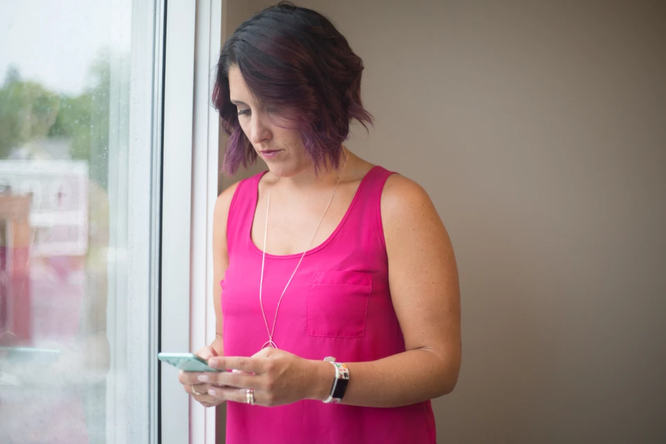 A woman standing beside a window looking down at a smartphone held in both hands