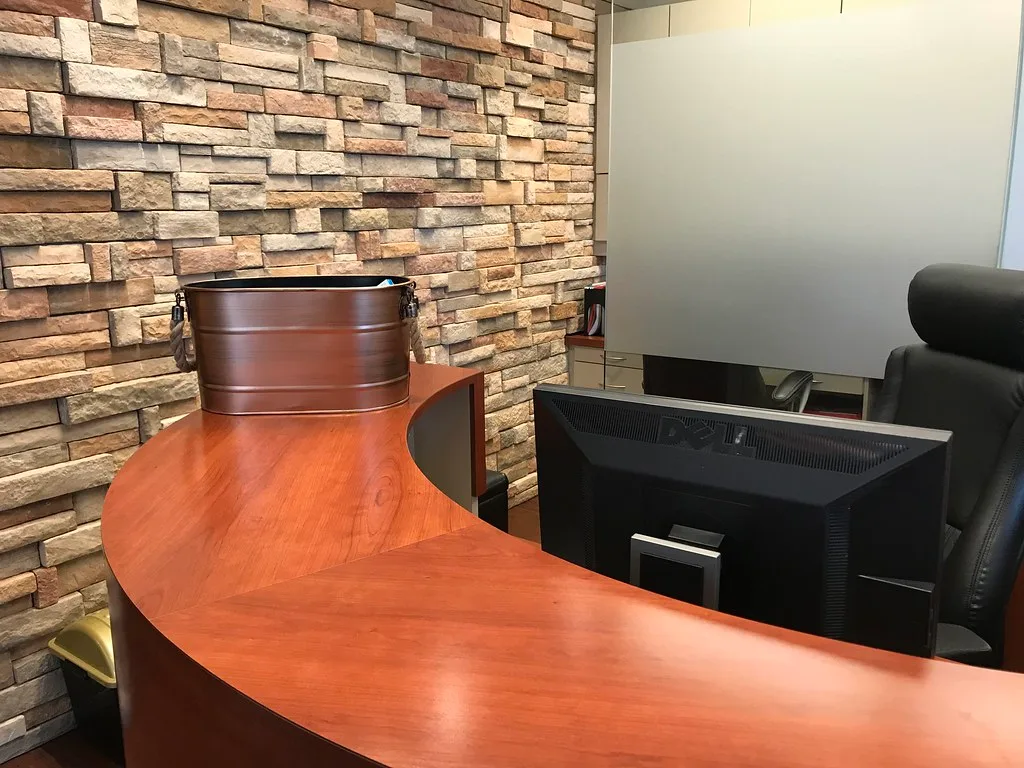 A curved wooden reception desk with a computer monitor and an office chair, against a stone veneer wall