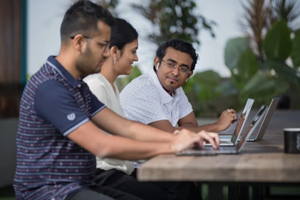 Three colleagues sitting side by side at a wooden table, each working on a laptop